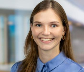 Portraitfoto einer jungen Frau. Sie hat braune Haare, trägt eine blaue Bluse und lächelt